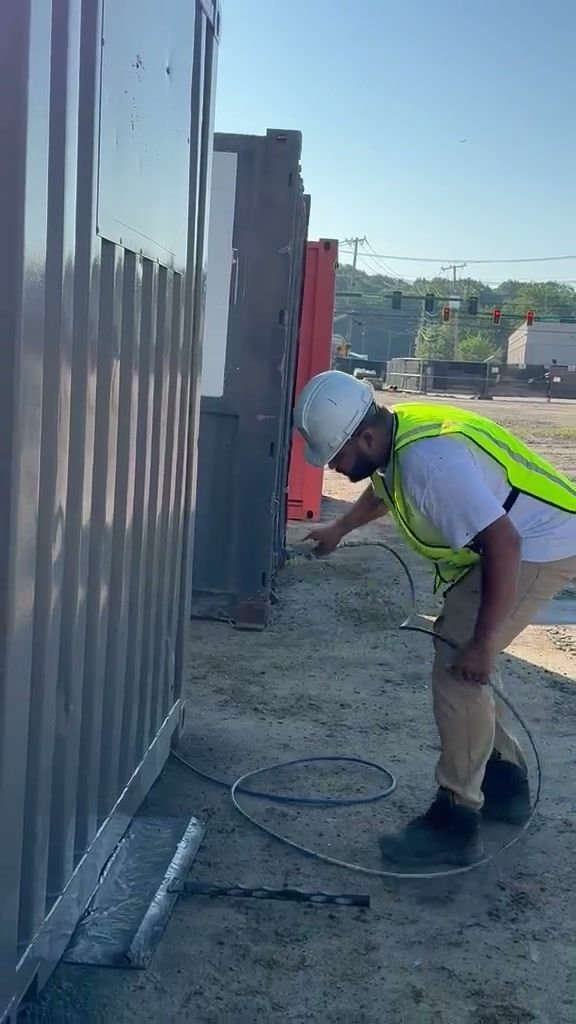 LNO Painting crew member in hard hat and safety vest spray-painting steel containers on a job site